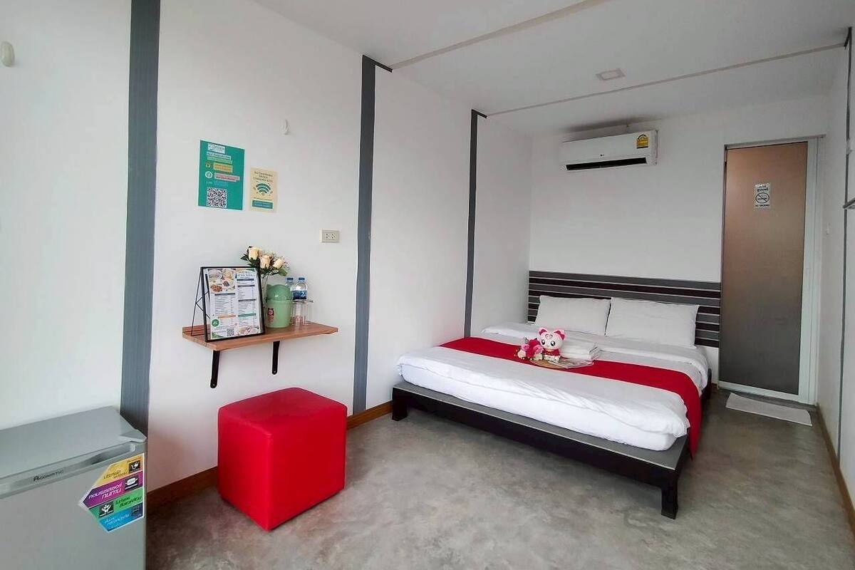 A hotel room with a bed, a red ottoman, a small shelf with amenities, and a door.