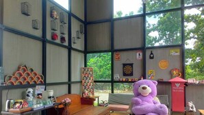 Una habitación con un ventanal grande, una mesa de madera, un sofá y un oso de peluche morado.