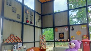 Una habitación con un ventanal grande, una mesa de madera, una silla, un osito de peluche y varias decoraciones en las paredes, incluyendo un dardo y un cartel de advertencia.