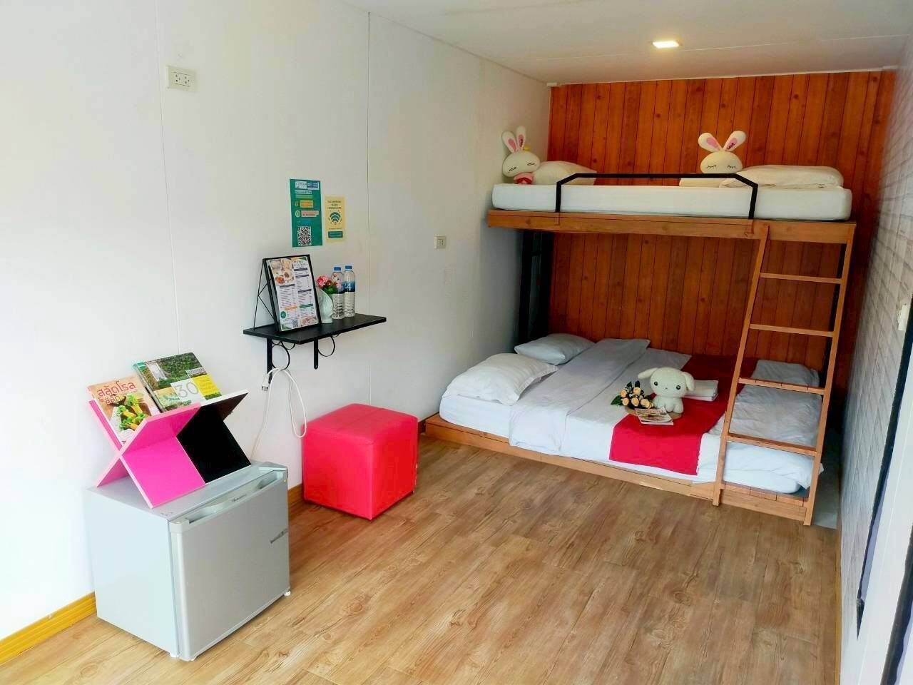 A compact room with a bunk bed, a small desk with a printer, a red ottoman, and a wooden wall panel.