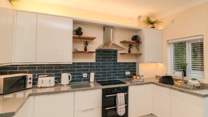 Une cuisine moderne avec des meubles blancs, un pare-feu noir en carreaux, un four encastré et une fenêtre avec des stores.