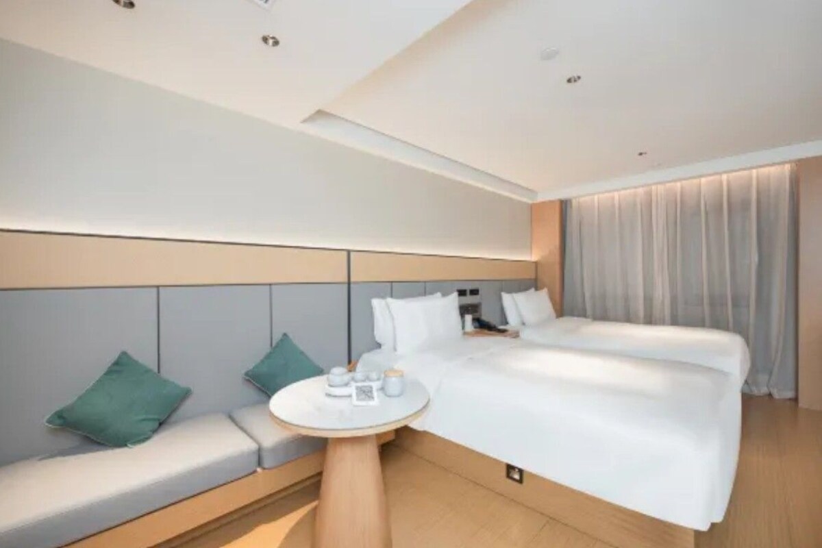 A modern hotel room with a large bed, a sofa, a small round table, and a headboard with built-in lighting.