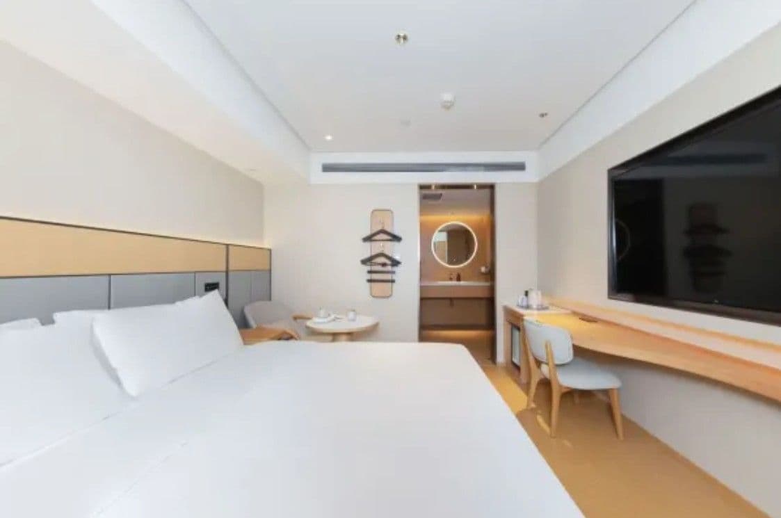 A modern hotel room with a large bed, a desk with two chairs, a round mirror, and a large window.