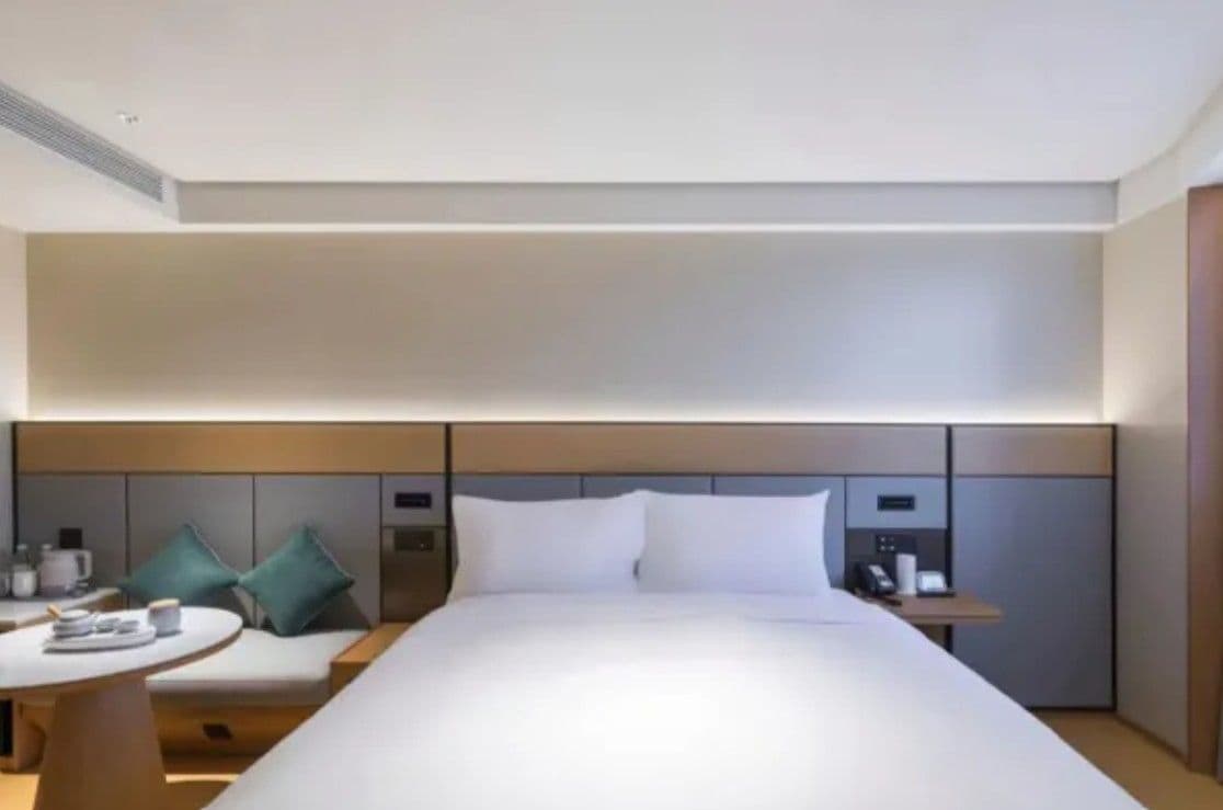 A modern hotel room with a large bed, a bedside table, a small round table, and a sofa with cushions.