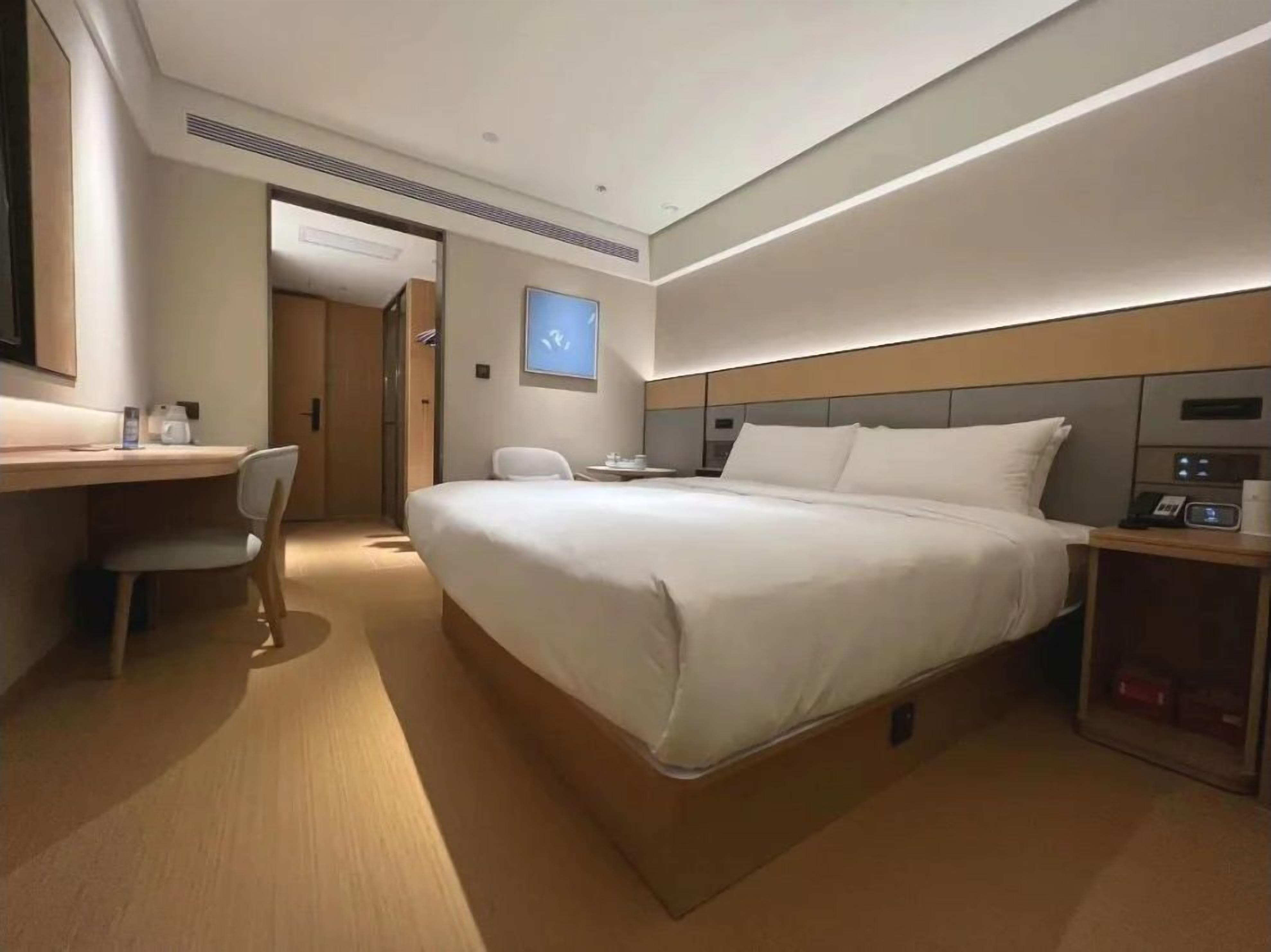 A modern hotel room with a large bed, a desk, a chair, and a small table with a phone and a lamp.