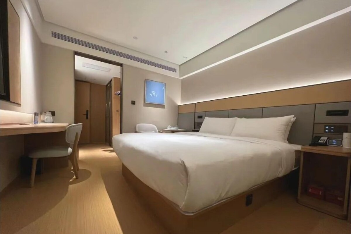 A modern hotel room with a large bed, a desk, a chair, and a small table with a phone and a lamp.