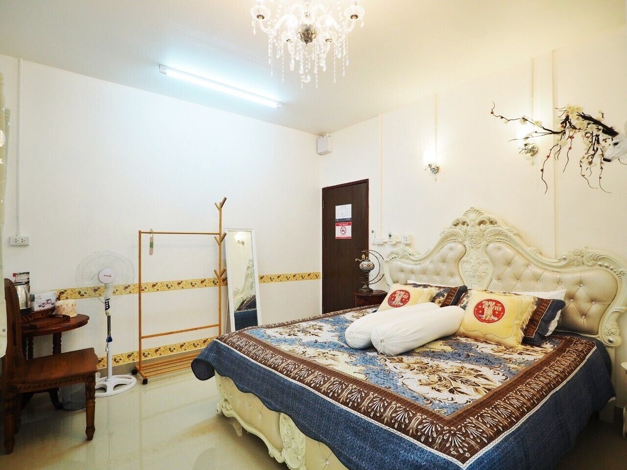 A bedroom with a large bed, a wooden chair, a standing fan, a mirror, and a chandelier.