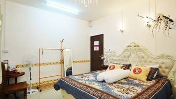 A bedroom with a large bed, a wooden chair, a standing fan, a mirror, and a chandelier.