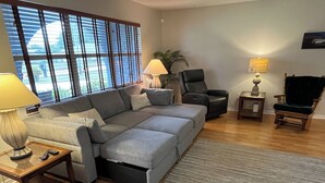 Ein modernes Wohnzimmer mit einem grauen Ecksofa, einem kleinen Holztisch, einem schwarzen Sessel, einem Holzhocker und einer Stehlampe.