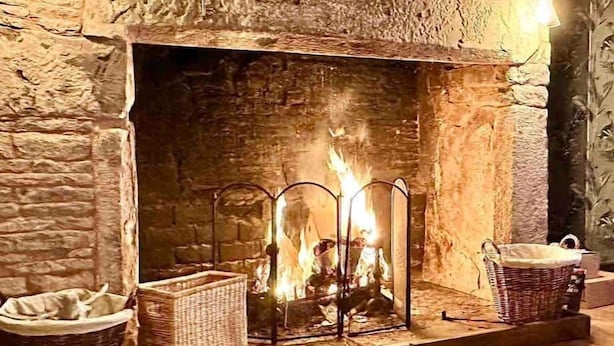 A stone fireplace with a fire, two baskets on either side, and a wooden bench in front. - 16th Cent,6 bed house,15 People (Monmouthshire)