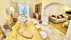 A spacious living area with a wooden dining table, cushioned seating, a chandelier, and a view through a glass door.