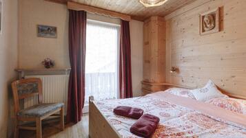 Una habitación de madera con una cama, una silla, un ventanal con cortinas y una lámpara fijada a la pared.