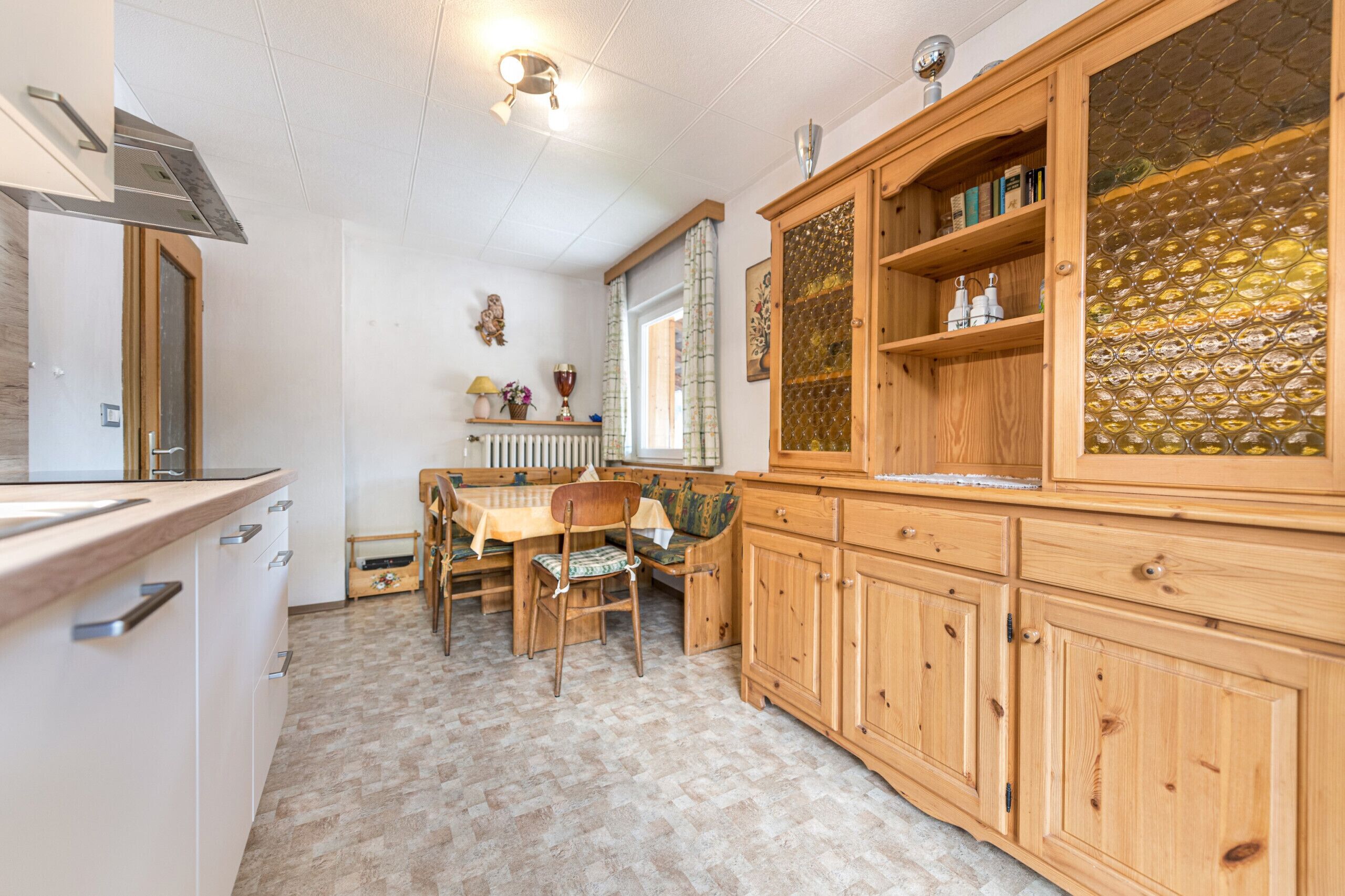 Une cuisine avec des meubles de rangement en bois, une table à manger, des chaises et une fenêtre avec des rideaux.