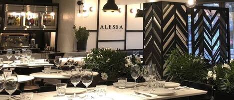 Un interior de restaurante con mesas dispuestas para comer, una zona de bar con botellas, y un letrero que dice "ALESSA".