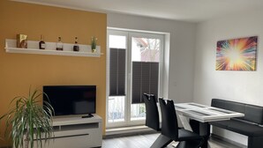 Un salon moderne avec un téléviseur, une table à manger, des chaises et un canapé. On y trouve une plante, une étagère avec des bouteilles et une œuvre d’art colorée accrochée au mur.