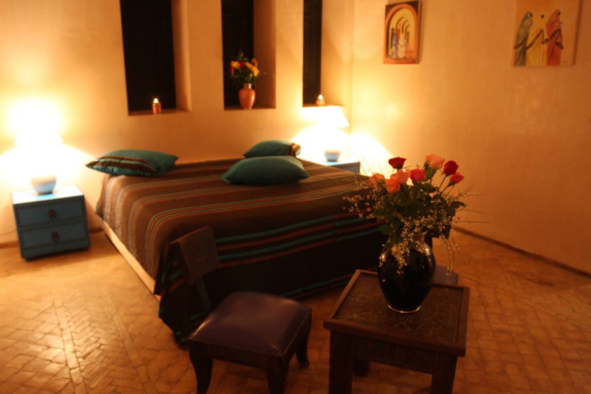 A hotel room with a bed, two bedside tables, a nightstand, a chair, a small table with a vase of flowers, and a wall with a painting.