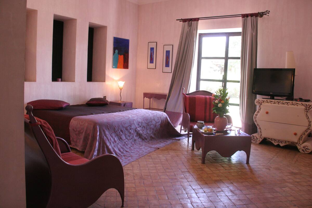 A hotel room with a bed, two chairs, a table with a vase, a television, and artwork on the walls.