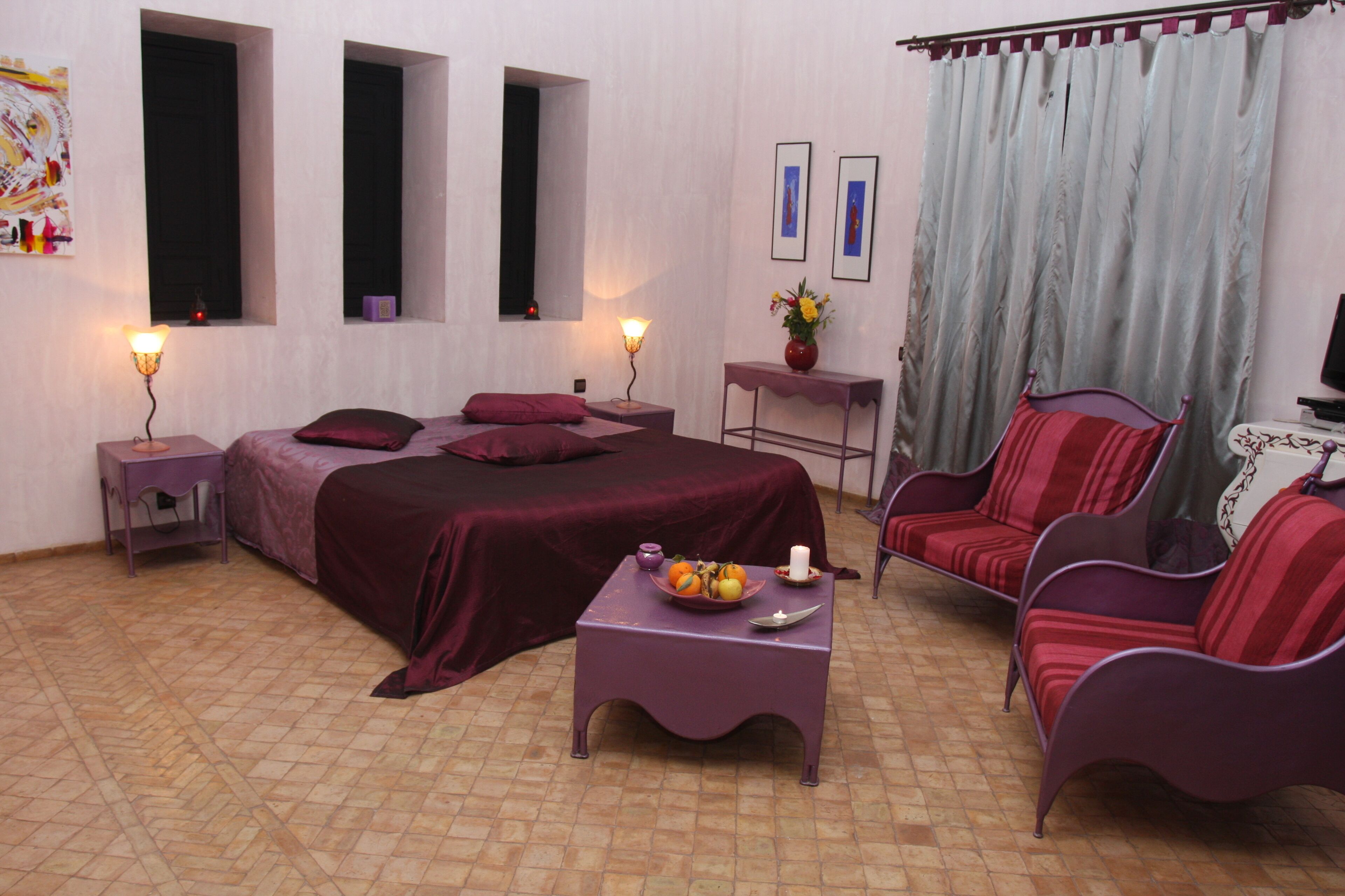 A bedroom with a large bed, two bedside tables, a nightstand, a chair, a small table with a vase of flowers, and a window with curtains.