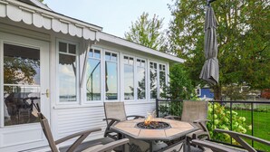 A covered patio with a fire pit table and chairs, large windows, and a view of a lake. - The Red Geranium Cottage of Big White Fish Lake! (Pierson Township)