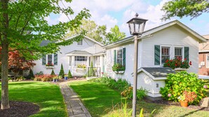 A white house with green shutters, a stone pathway, and a garden with various plants and flowers. - The Red Geranium Cottage of Big White Fish Lake! (Pierson Township)