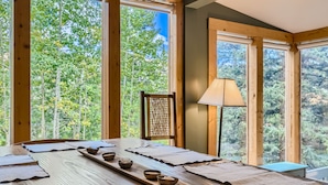 Una mesa de madera para comer con individuales y cuencos, frente a grandes ventanales con vistas a un bosque.