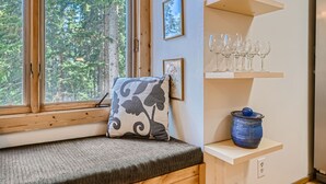 Habitación acogedora con un banco junto a una ventana que da a un bosque, una estantería con copas de vino y una maceta azul sobre una mesita.