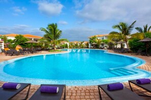 A swimming pool with lounge chairs and umbrellas, surrounded by palm trees and buildings. - VILLA TORTUGA BEACH RESORT (Sal)