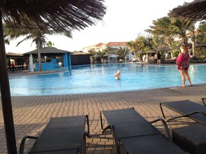 A swimming pool with lounge chairs and umbrellas, surrounded by palm trees and a building with a red roof. - VILLA TORTUGA BEACH RESORT (Sal)