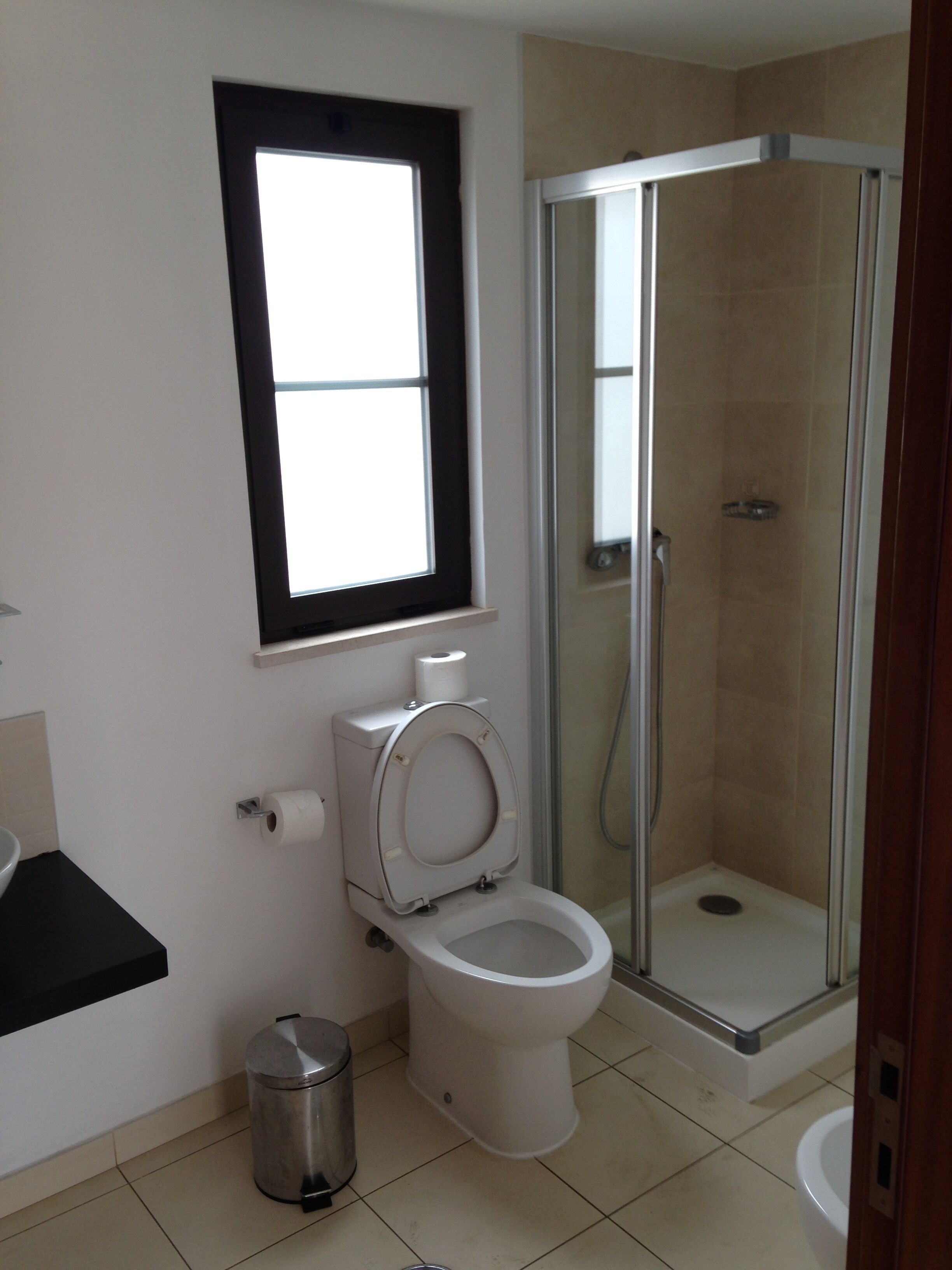 A bathroom with a toilet, a trash can, a window, and a glass-enclosed shower.