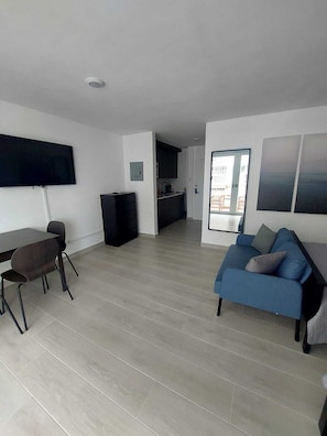 Una sala de estar moderna con un televisor de pantalla plana, un comedor con mesa y sillas, un sofá azul y una cocina al fondo.