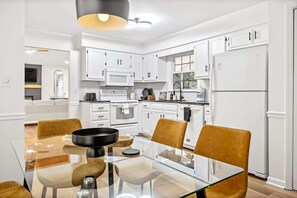 A modern kitchen with white cabinets, a glass dining table, and a black hanging light fixture.