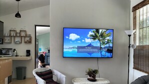 A living room with a flat-screen TV mounted on the wall displaying a tropical beach scene. There is a wicker chair with a striped cushion, a small round table with a plant, and a grey sofa. A kitchen area with a microwave and coffee maker is visible in the background.