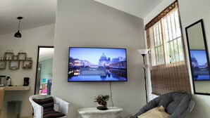 A modern living room with a flat-screen TV mounted on the wall displaying a cityscape. There is a wicker chair, a small round table with a plant, and a grey sofa with a cushion.