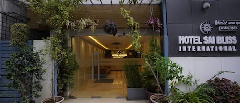 The entrance of Hotel Sai Bliss International with a modern design, featuring a glass door, potted plants, and a signboard.