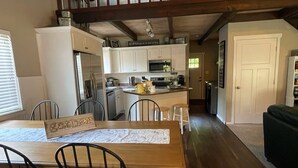 Un coin repas avec une table et des chaises en bois, une cuisine à l’arrière-plan et un escalier menant à un niveau supérieur.