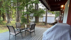 Une terrasse extérieure couverte, avec une table et des chaises, entourée d’arbres et offrant une vue sur une maison au loin.