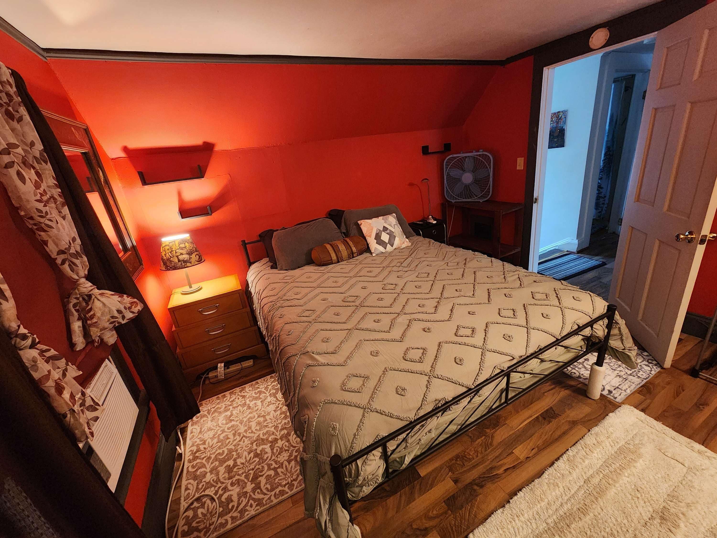 A bedroom with a bed, bedside tables, a lamp, a fan, and a door leading to another room.