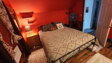 A bedroom with a bed, bedside tables, a lamp, a fan, and a door leading to another room.