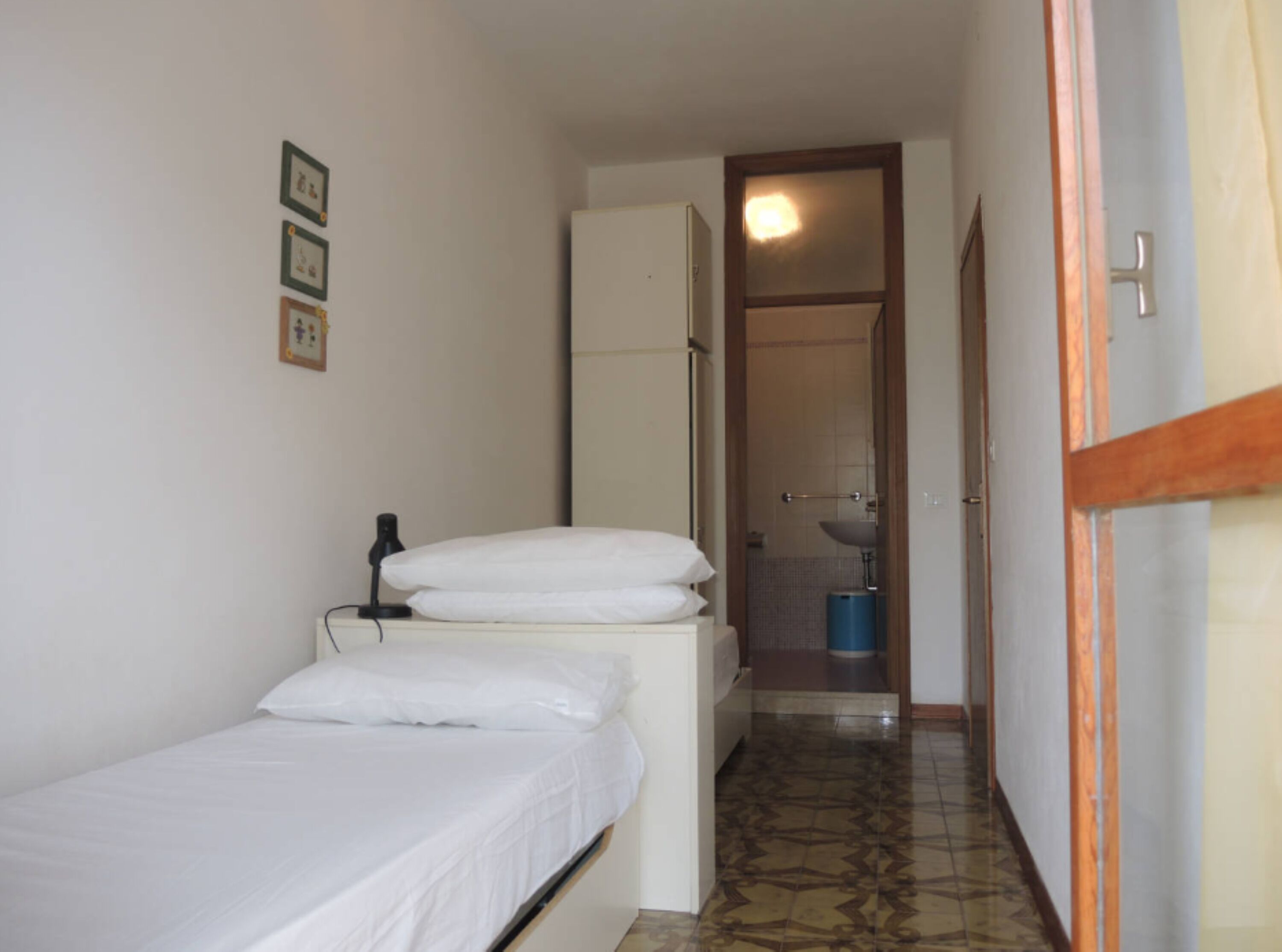 Ein schmaler Flur mit einem Bett links, einem weißen Schrank und einem Badezimmer im Hintergrund.