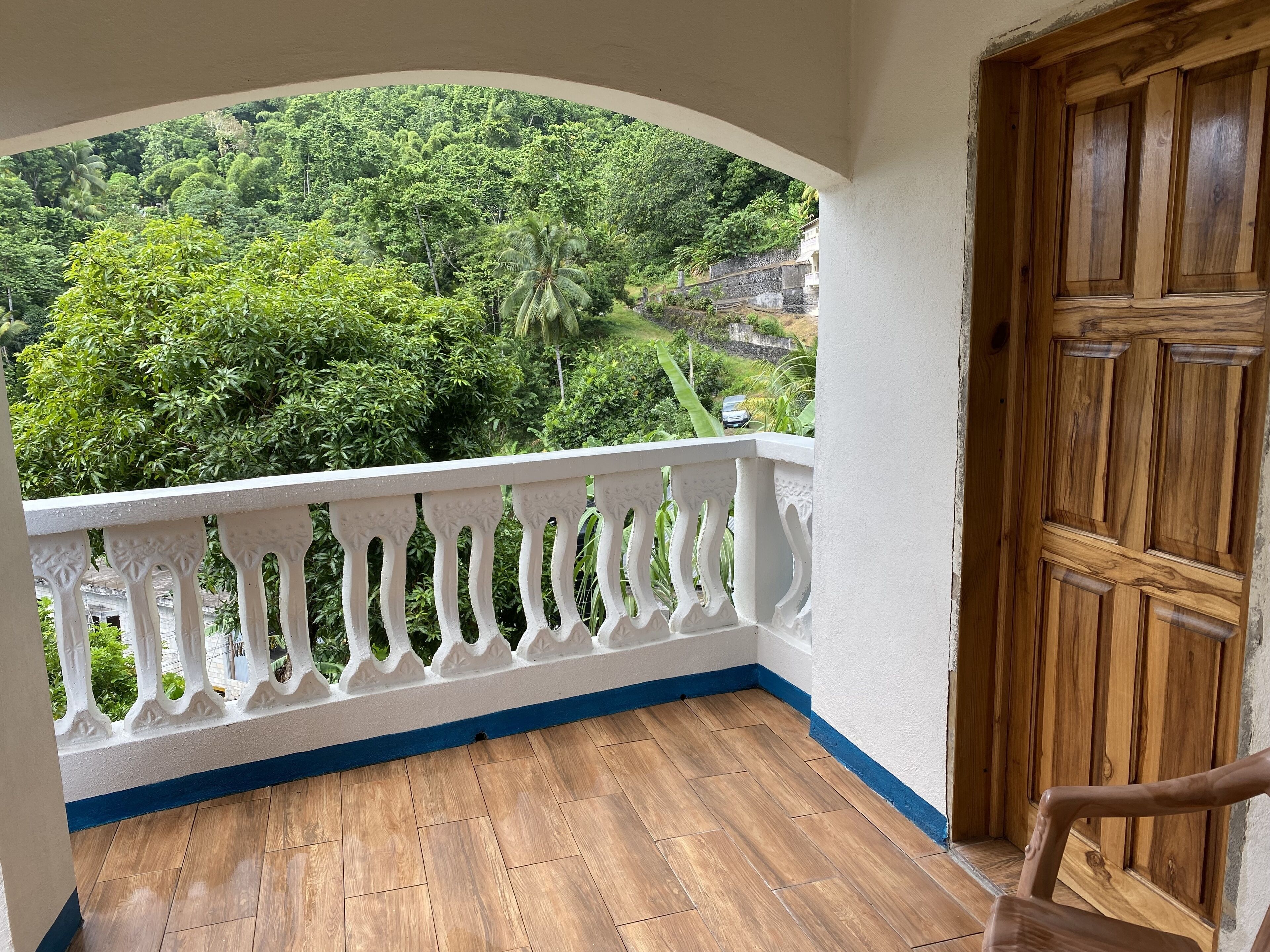 A balcony with a wooden floor, white railing, and a view of lush greenery and a stone wall.