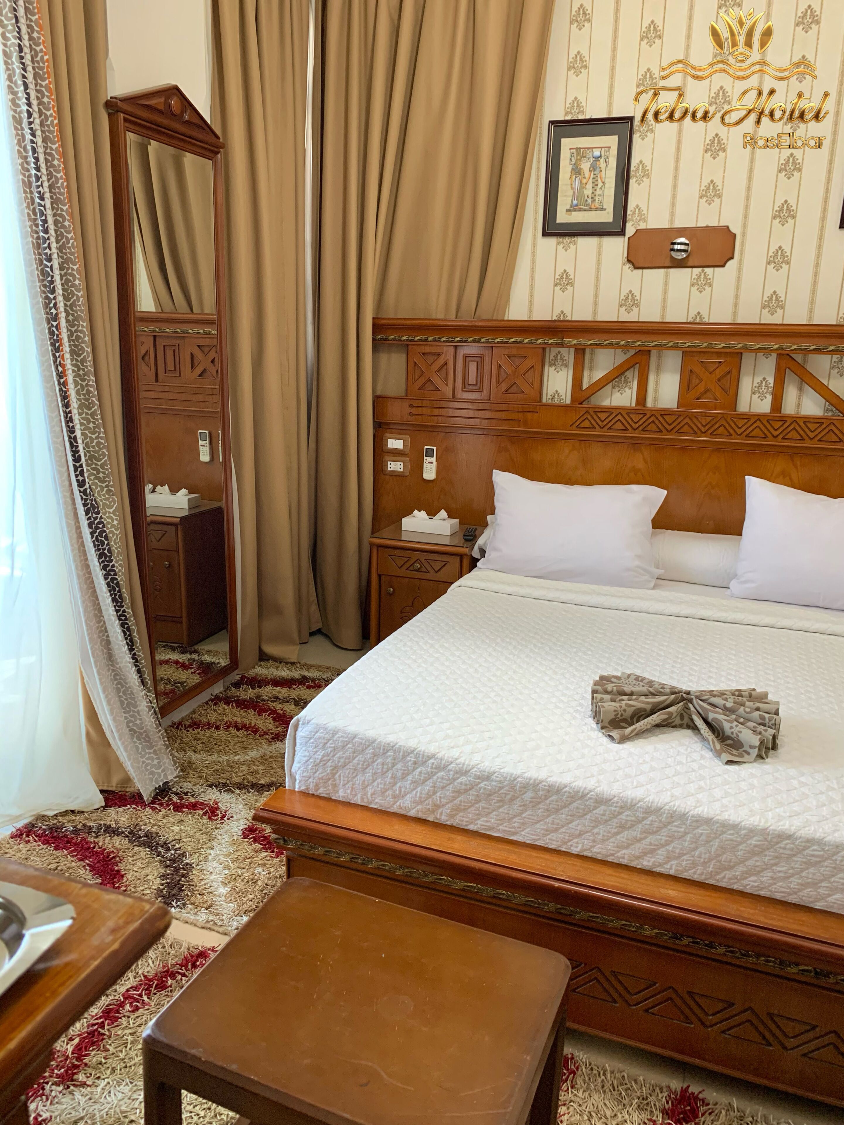 Habitación de hotel con una cama grande, cabecera de madera, mesita de noche con lámpara, espejo y armario.