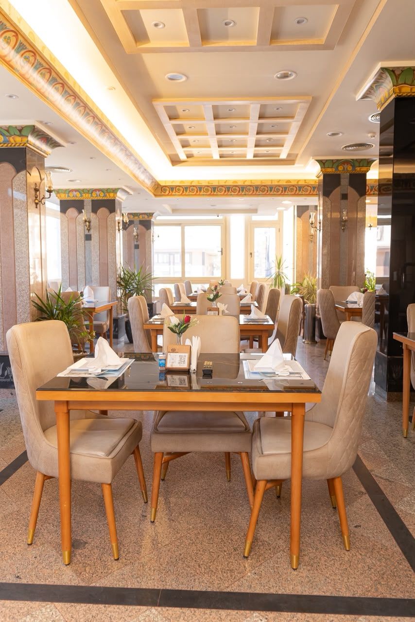 A hotel dining area with tables and chairs, decorative ceiling, and large windows.