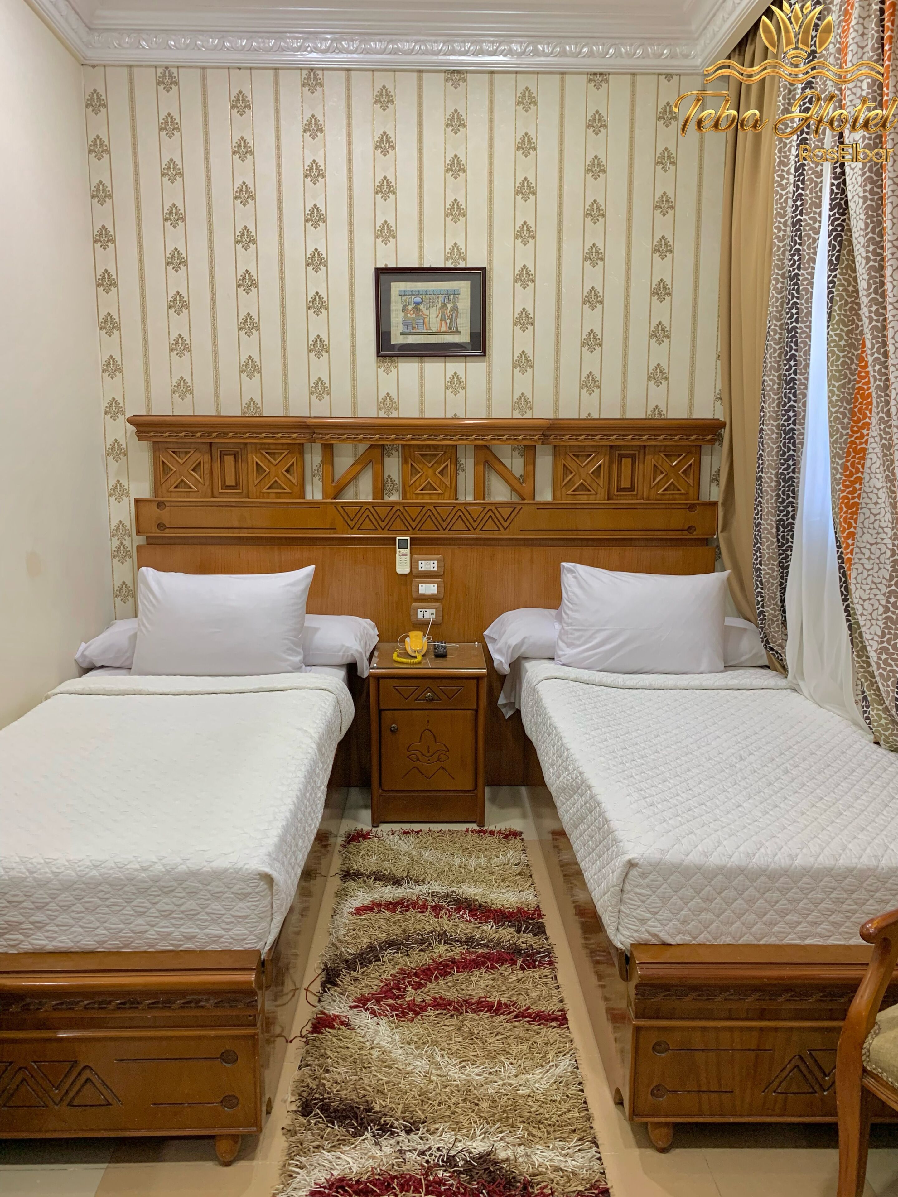 Ein Hotelzimmer mit zwei Betten, einem Holzkopfteil, einem Nachttisch mit Telefon und einem gemusterten Teppich.