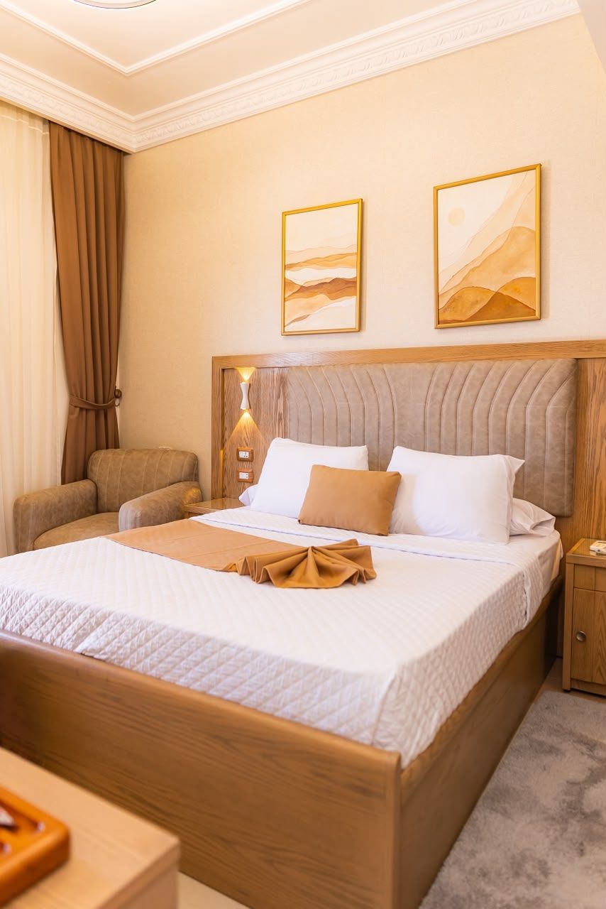 Una habitación de hotel con una cama grande, cabecera de madera, mesita de noche, una silla y dos cuadros en la pared.
