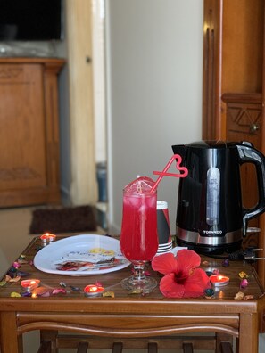 Une table avec un verre d’une boisson rouge, une cafetière noire, une assiette contenant des restes de nourriture et une fleur rouge.