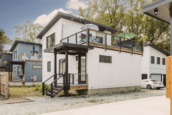 A modern two-story house with a balcony, a small outdoor seating area, and a car parked in the driveway.