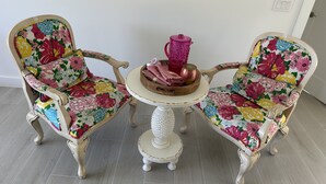 Zwei Stühle im Vintage-Stil mit floralem Bezug, ein kleiner runder Tisch mit Zierfuß und eine rosa Karaffe darauf.