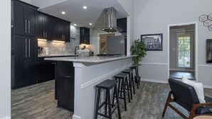 A modern kitchen with dark cabinets, a central island with stools, and a dining area with a chair and table. - 4 Bedroom Retreat — Close to Ft Benning: Modern, Cozy, and Mins to Everything! (Columbus)