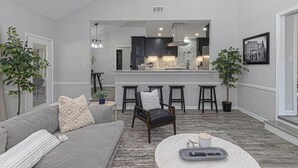 A modern living room with a grey sofa, a coffee table, and a kitchen area in the background. - 4 Bedroom Retreat — Close to Ft Benning: Modern, Cozy, and Mins to Everything! (Columbus)