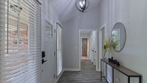 A hallway with a wooden floor, white walls, and a console table with a mirror and vases. - 4 Bedroom Retreat — Close to Ft Benning: Modern, Cozy, and Mins to Everything! (Columbus)